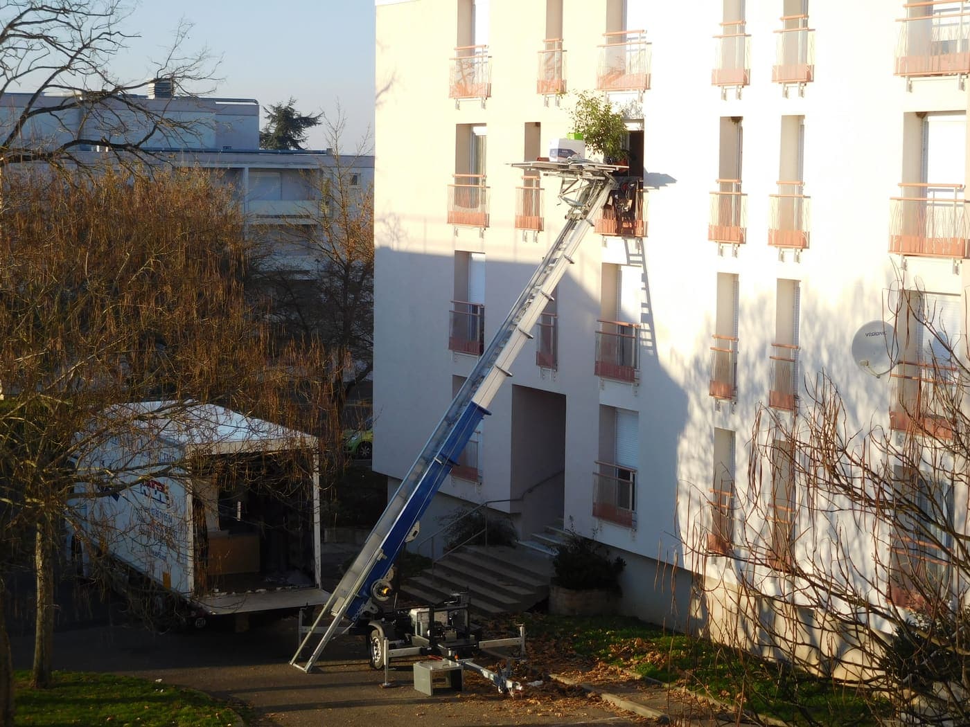 Une remorque monte-meuble posée au pied d'un immeuble d'habitation : l'échelle inclinée monte depuis la remorque, calée sur ses béquilles, jusqu'à une fenêtre d'étage, où le plateau est arrêté avec des objets posés dessus. Le camion de déménagement est garé plus loin, sur l'allée.
